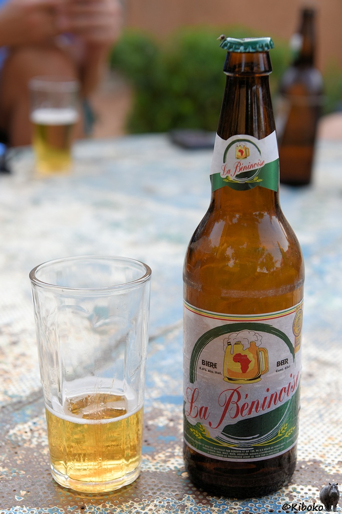 Das Bild zeigt eine Bieflasche mit weiß-grünem Ettikett mit roter Schrift: La Béninoise. Daneben steht ein fast leeres Glas auf einem rostigen Tisch aus Riffelblech.