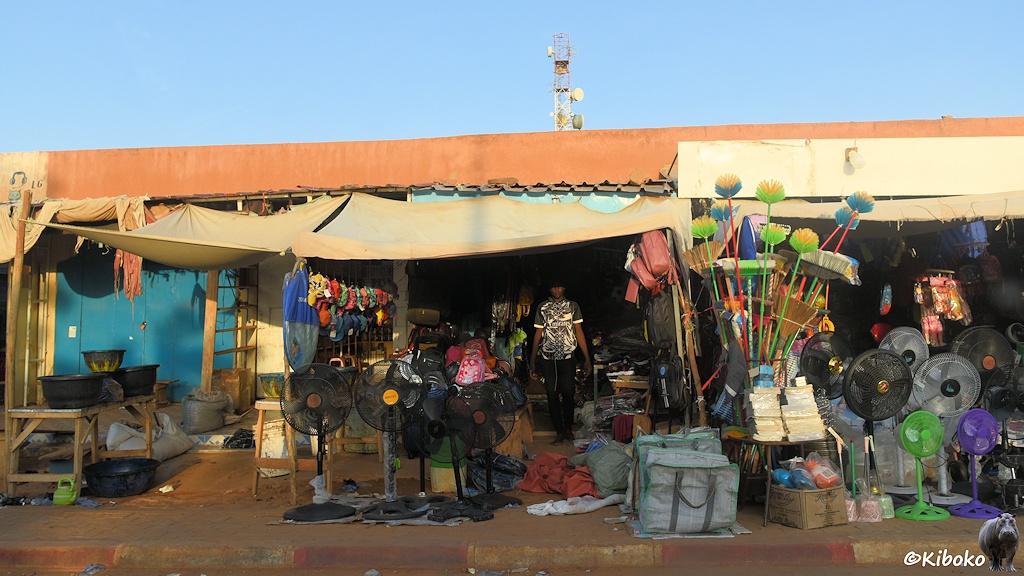 Das Bild zeigt mehrere Geschäfte einer Geschäftsstraße in Garagengröße. Auf dem Gewhweg stehen Standventilatoren und große Taschen. An der Ecke des Geschäfts steht ein Straus von bunten Plastikbesen mit dem Stil nach unten.