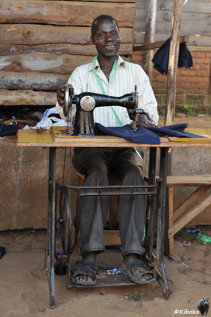 Das Bild zeigt einen sitzenden Mann hinter einer schwarzen Nähmaschine im Hochformat. Der Mann lächelt und hat ein grünes Maßband um den Hals gelegt. Mit den Händen führt er einen blauen Stoff an die Nähmaschine. Mit den Füßen treibt er die Maschine an.