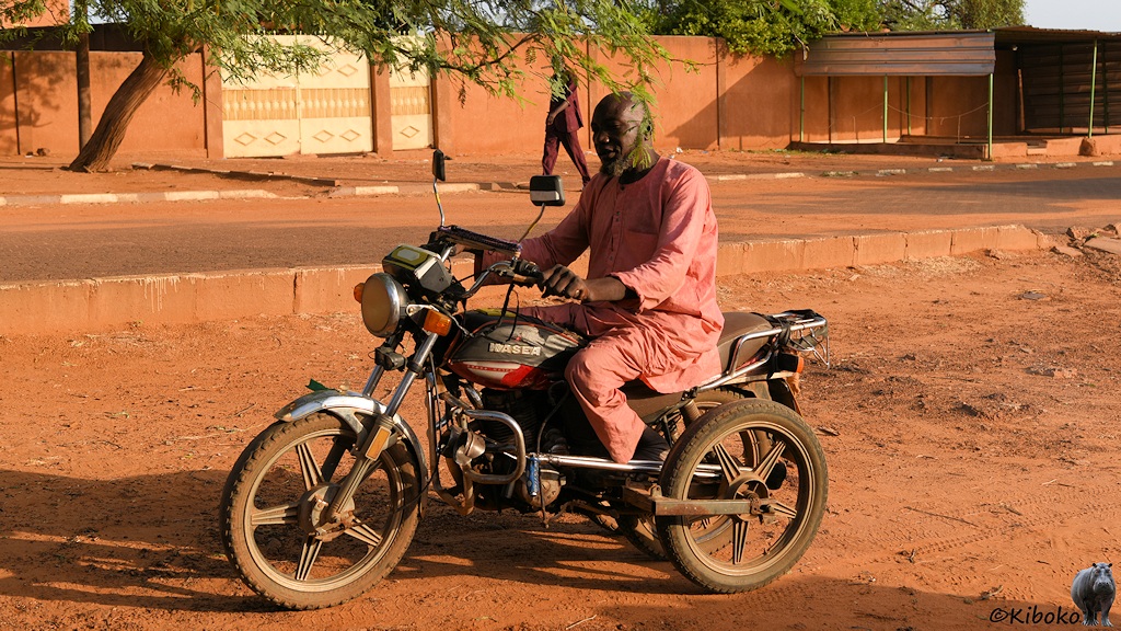 Das Bild zeigt einen älteren Mann in einem Rosa gewandt auf einem dunkelgrauen Motorrad. Hinten hat das Motorrad drei gleichgroße Räder.