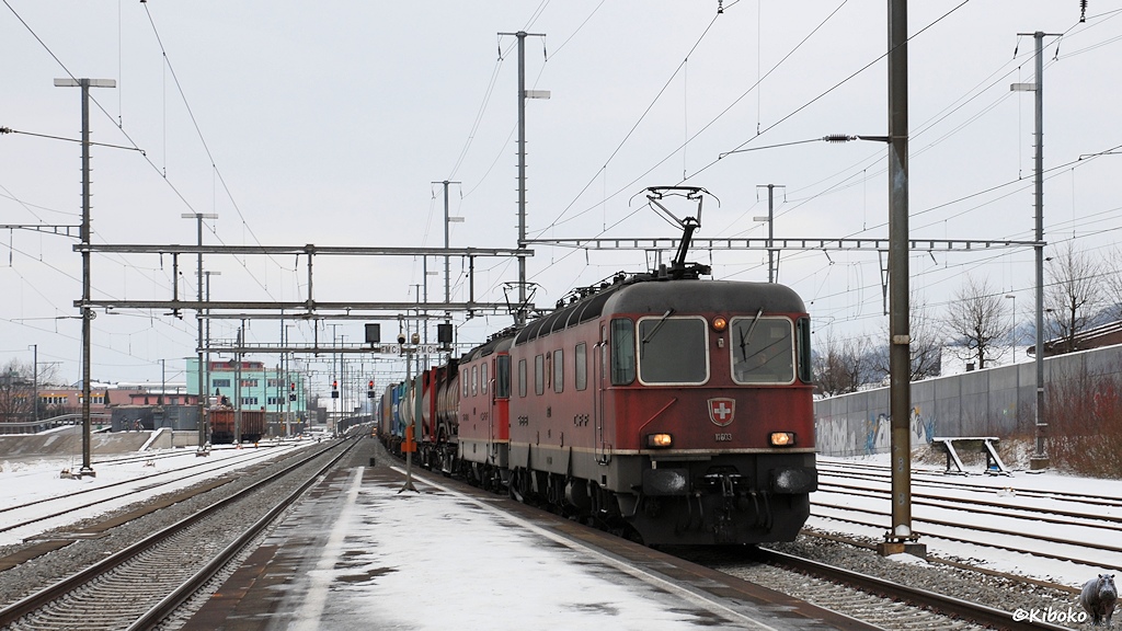 Das Bild zeigt zwei rote Elektrolokomotiven mit einem Containerzug bei der Durchfahrt durch einen Bahnhof.