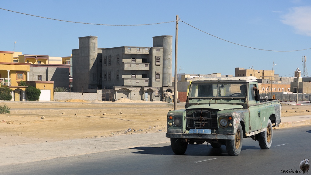 Das Bild zeigt einen ausgeblichenen grünen Land Rover mit Pritsche. Es ist ein sehr altes Modell. Im Hintergrund ist ein neues, betongraues, dreigeschossiges Gebäude mit halbrunden Türmen.