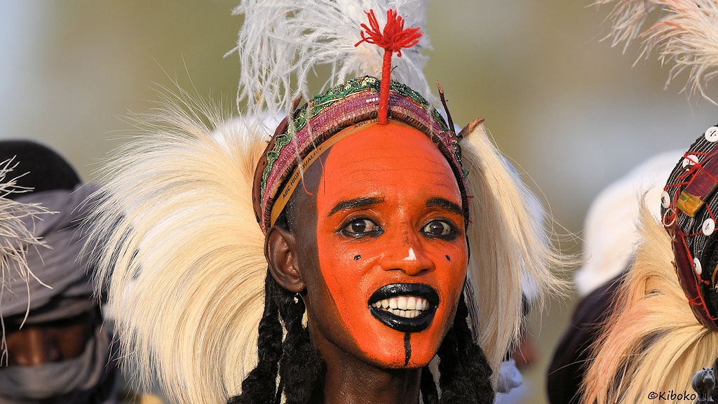 Das Bild zeigt das Porträt eines Mannes mit orange gefärbtem Gesicht, schwarzen Lippen, schwarzen Augenlieder mit geöffnetem Mund und Augen.