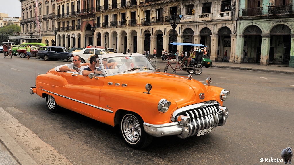 Das Bild zeigt ein altes oranges Cabrio amerikanischer Bauart. Das Verdeck ist offen. Im Auto sitzen neben dem Fahrer ein Mann, sowie zwei Frauen und ein Mann auf der Rückbank. Im Hintergrund ist ein mehrstöckiges, heruntergeommenes Gebäude mit Arkardengang.