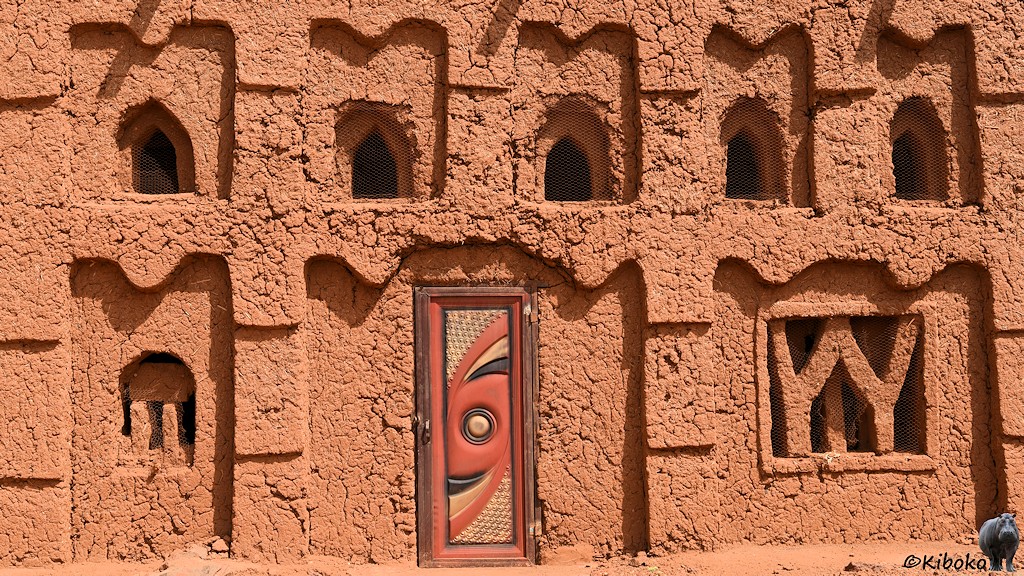 Das Bild zeigt einen Ausschnitt der Lehmmoschee mit einer Holztür in der Mitte. Die Tür hat in der Mitte einen Kreis darum ist eine Spriale aus rotem, schwarzem und beigem Streifen. Die Ornamente an den Seiten haben drei Stufen. Darüber ist eine Welle. Der Lehm ist sehr rissig mit tiefen Furchen.