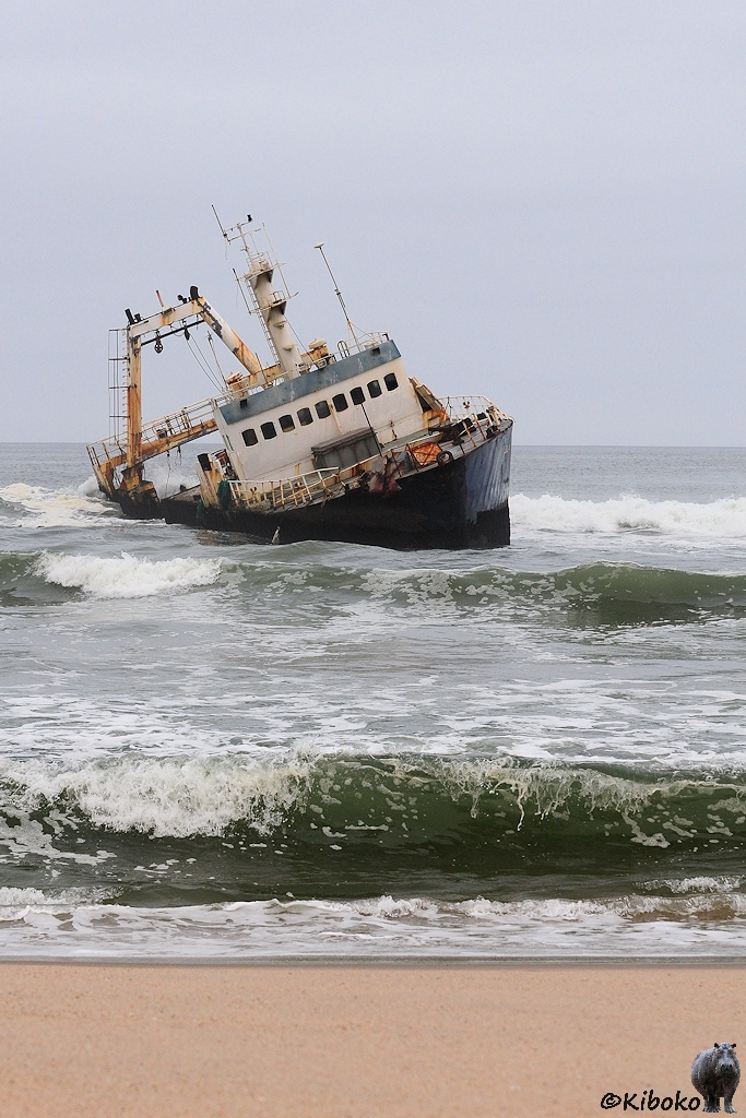 Hochformataufnahme vom Schiff im Schräglage mit Wellen und Strand im Vordergrund.