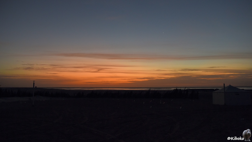 Das Bild zeigt eine Morgenstimmung. Der Vordergrund ist noch dunkel. Am rechten Bildrand ist eine viereckige Hütte. Am linken Bildrand ist ein Turm am Horizont. An der Horizotnlinie ist ein Wasserstreifen. Über dem Horizont ist einer oranger Streifen mit Wolken am Himmel darüber geht der Himmel über hellblau in ein tiefes Dunkelblau über. Darin leuchtet die Venus als letzter Stern.