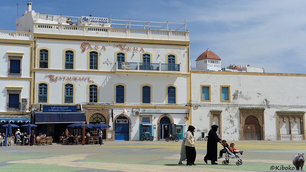 Das Bild zeigt ein dreigeschossiges, weißes Gebäude mit blauen Türen und Fenstern an einem Platz. Die Gebäudekanten und Fenster sind hellbraun eingefasst. Im linken Teil des Gebäudes ist ein Restaurant. Auf dem Platz laufen im Vordergrund drei verschleierte Frauen mit einem Kinderwagen, auf dem zwei Kinder sitzen.