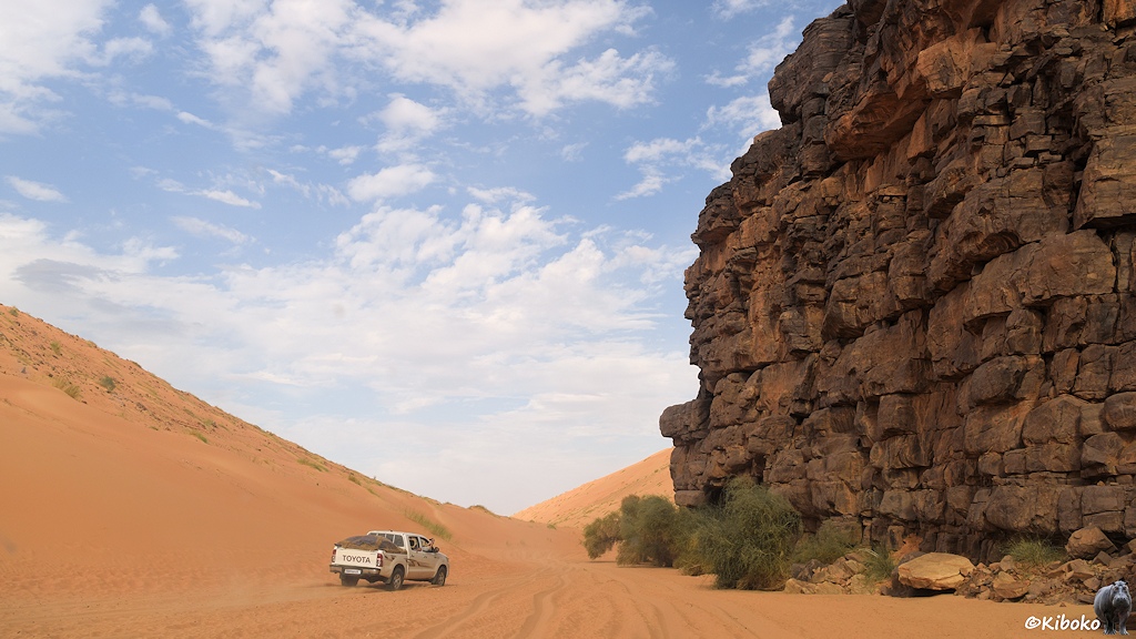 Das Bild zeigt einen vorausfahrenden weißen Geländewagen im lachsfarbenen Tiefsand in einem engen Tal. Rechts ist eine senkrechte Felswand aus dunklen Steinen. Rechts ist ein steiler Hang aus lachsfarbenen Sand.