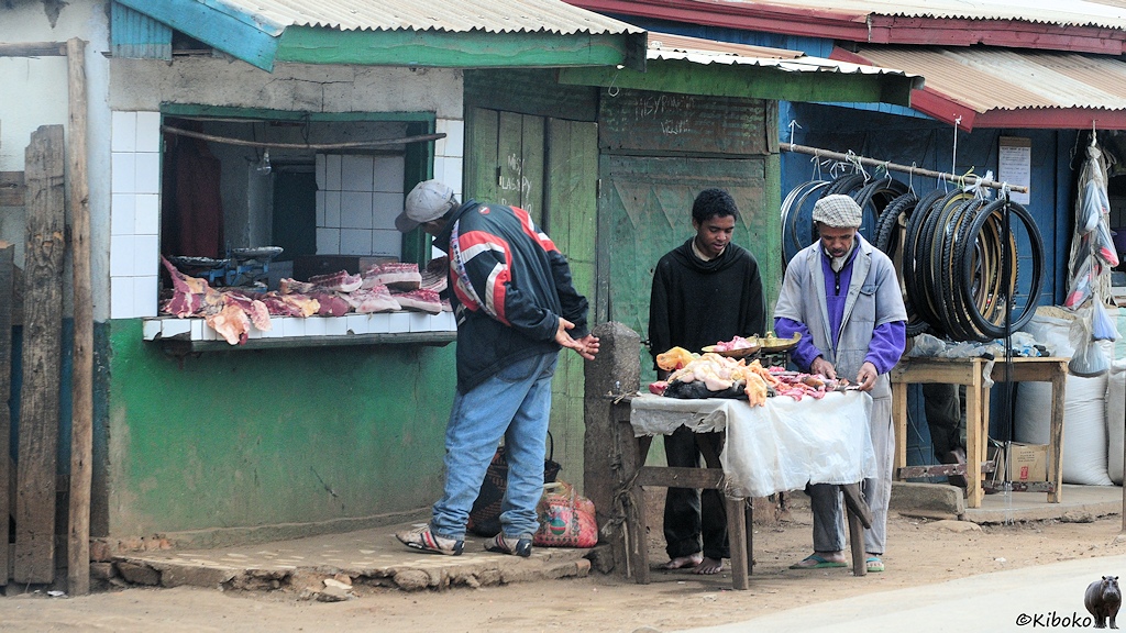 Das Bild zeigt eine ein kleines weiß-grünes Haus mit großem, glaslosen Fenster. In der Fensterbank liegen Fleischbrocken. Ein Mann beugt sich zum Fleisch. Vor dem Haus steht ein zusammengezimmerter Holztisch mit weiteren Fleischstücken auf einer ausgefransten weißen Decke. Zwei Männer schauen sich das Fleisch an. Der Boden ist staubig. Im Hintergrund hängen Fahrradreifen vom nächsten Geschäft.