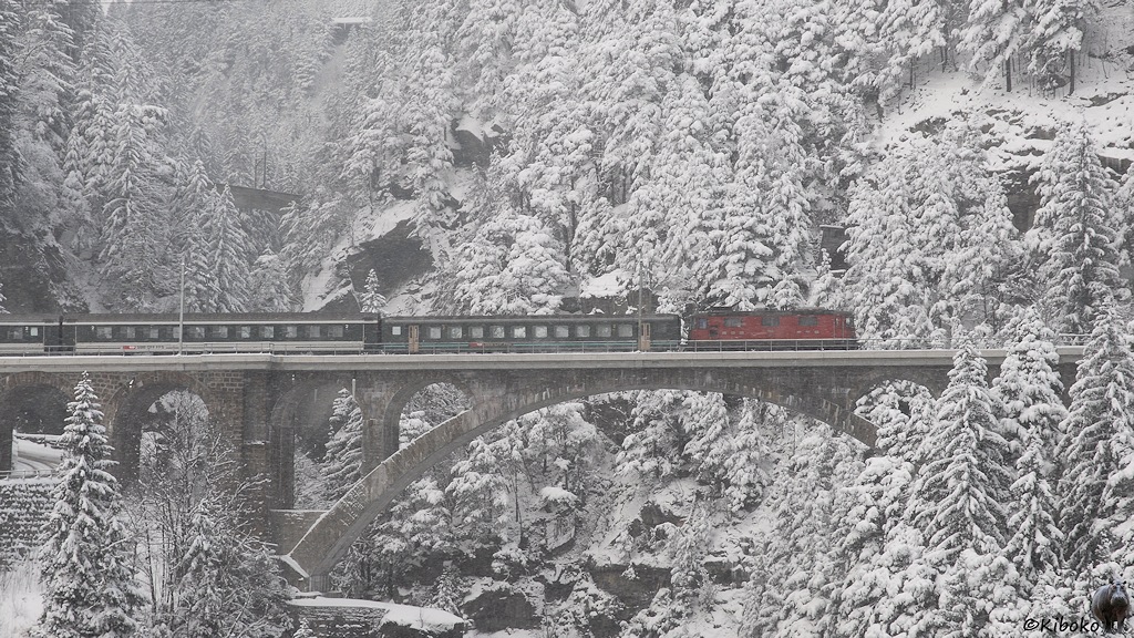Das Bild zeigt eine rote Elektrolokomotive mit einem grünen und zwei grün-weißen Personenwagen auf einer großen Steinbogenbrücke. Drumherum stehen Nadelbäume im Schneemantel.