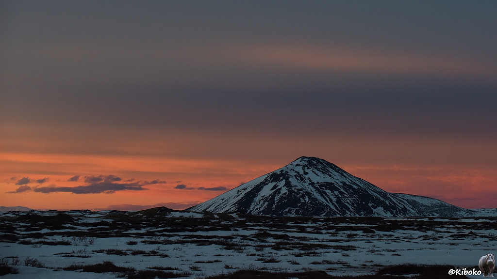 Ein spitzer Berg steht auf einer schwarzen Ebene mit Schneeflecken. Hinter dem Berg leuchtet der Himmel orange.