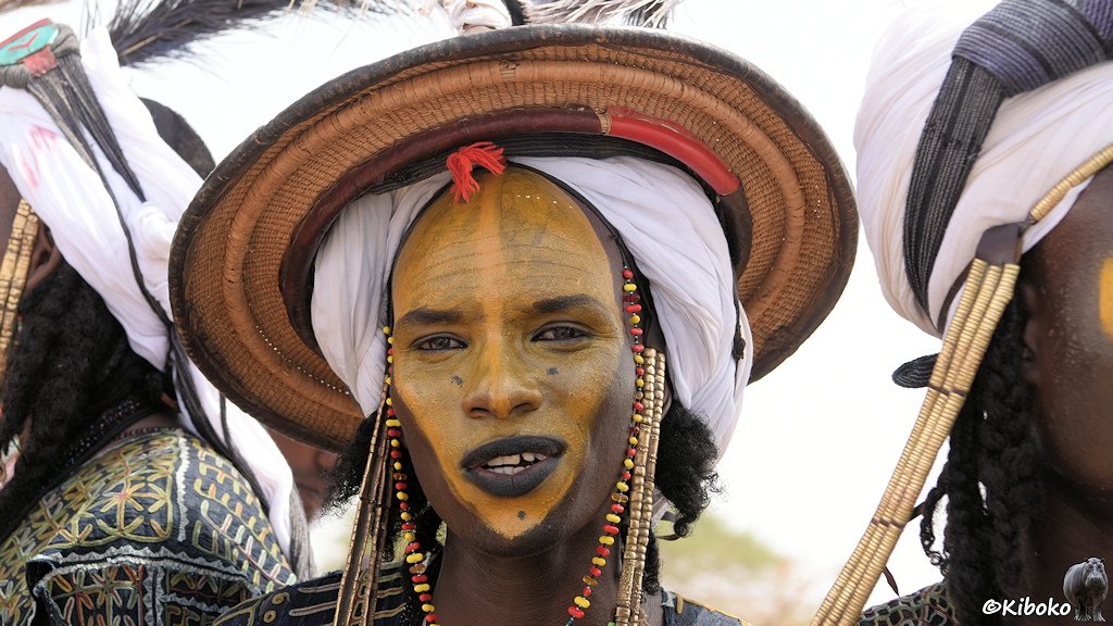 Das Bild zeigt das Porträt eines Mannes mit gelb eingefärbtem Gesicht und schwarzen Lippen. Er trägt einen runden Strohhut über einen weißen Turban.