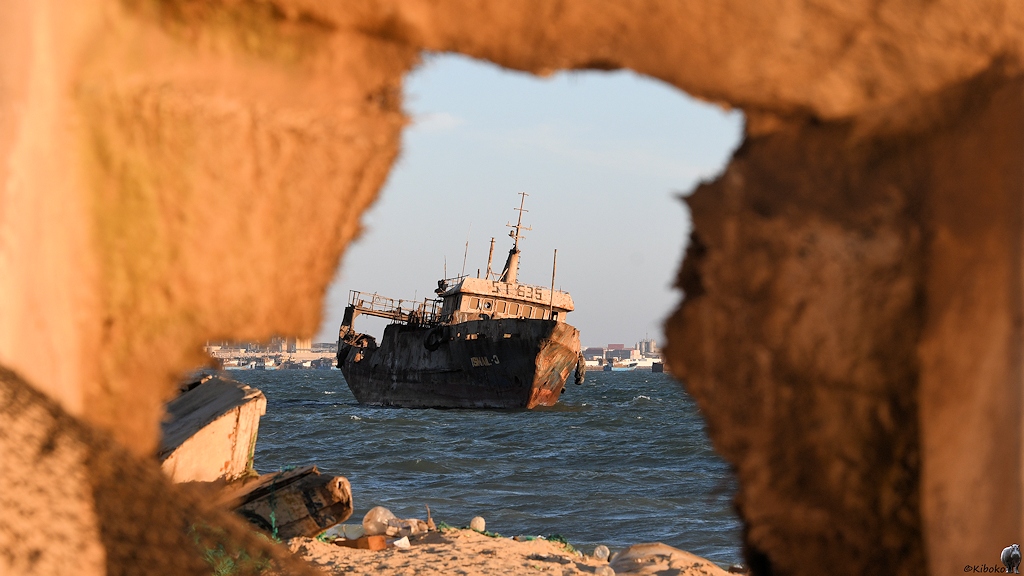 Das Bild zeigt den Blick durch ein Loch in einem Beiboot aus Kunststoff. Dahinter ist ein Fischtrawler mit blau-roten Rumpf und weißen Aufbauten.