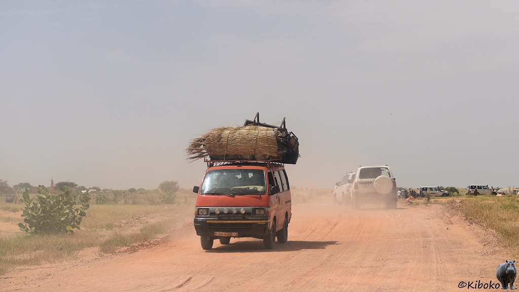 Das Bild zeigt einen roten Kleinbus mit vollbeladenen Dachgeäckträger auf einer staubigen Pista us lachsfarbenen Sand. Mehrere weiße Geländewagen stehen am rechten Straßenrand.