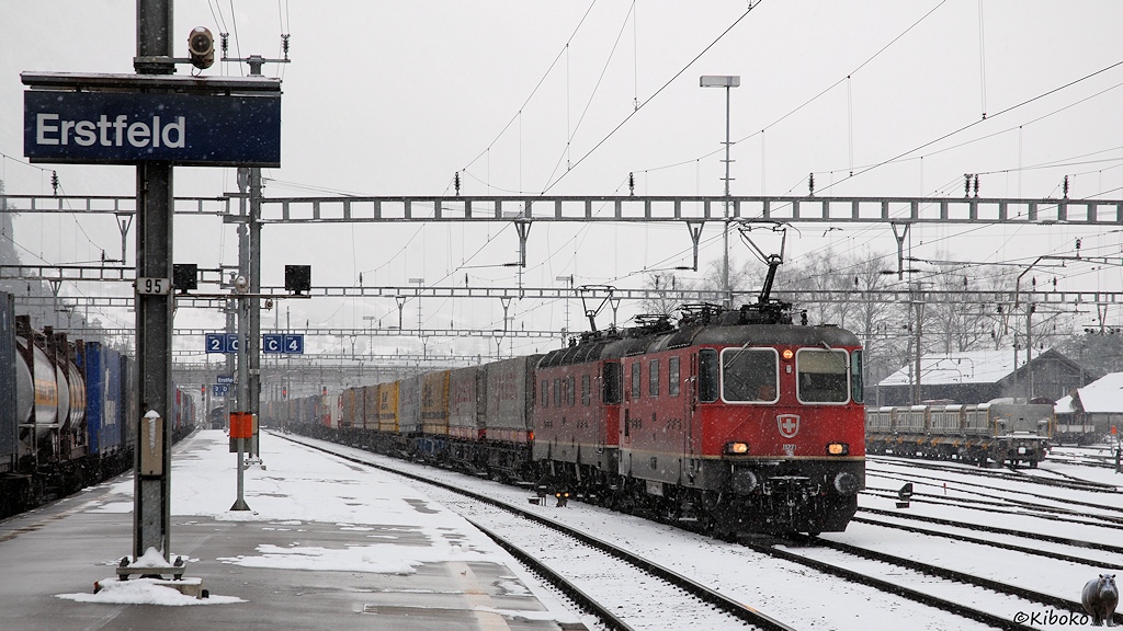 Das Bild zeigt zwei rote Elektrolokomotiven vor einem Güterzug mit LKW Aufliegern bei der Fahrt durch einen verschneiten Bahnhof. Links steht das Bahnhofsschild mit der Aufschrift Erstfeld.