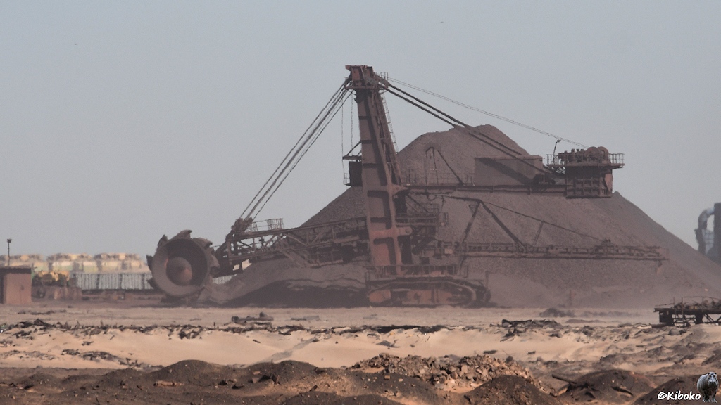 Das Bild zeigt einen rotbraunen Schaufelradbagger. Das Schaufelrad hat einen Durchmesser von ca. 10m. Es hat 9 Schaufeln. Der Bagger steht auf einen Kettenfahrwerk. Der Bagger ist über 30m hoch. Dahinter ist ein noch größerer Berg aus rotbraunem Eisenerz.