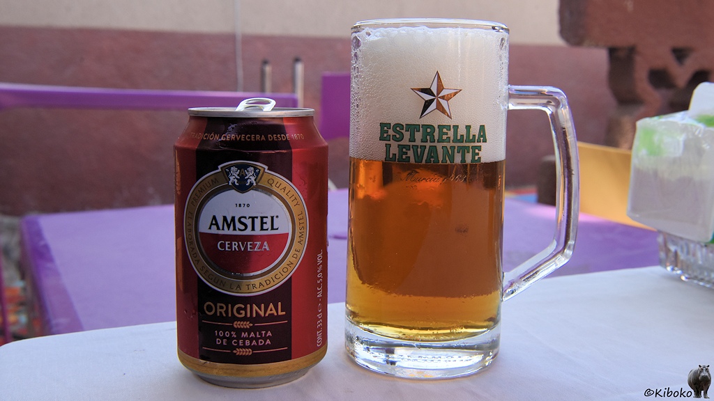Das Bild zeigt eine geöffnete rote Bierdose mit mit einem weiß-roten, runden Emblem, das von einen goldenen Ring umgeben ist. Darin steht AMSTEL CERVEZA. Unter dem Emblem steht mit Goldener Schrift: ORIGINAL. Darunter steht zwischen zwei waagerechten Ähren: 100% MALTA DE CEBADA. Daneben steht gefülltes Bierglas mit Henkel. Das obere Drittel ist mit Schaum gefüllt. Vor dem Schaum steht auf dem Glas: Estrella Levante und ein fünfeckiger Stern mit Diamantenschliff.
