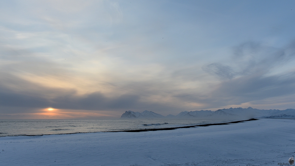 Das Bild zeigt eine Landschaftsaufnahme mit einer weiten Bucht. Der Strand ist mit Schnee bedeckt. Zwischen Wasser und Schnee ist ein schwarzer Sandstreifen. Am Ende der Bucht ist eine Bergkette. Über dem Meer taucht die tiefstehende Sonne unter einer Wolkenschicht wieder auf.