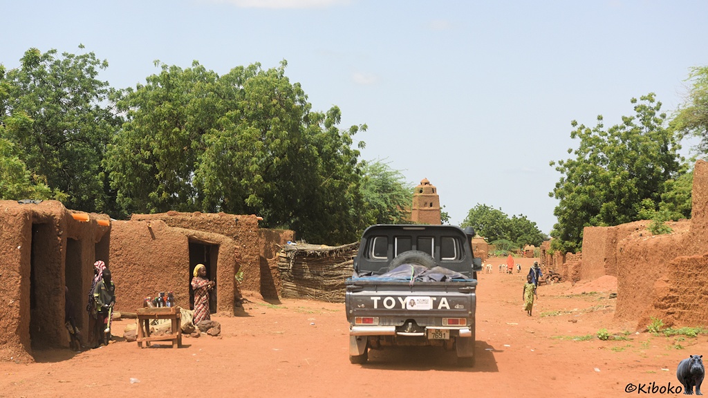 Das Bild zeigt einen grünen Geländewagen von hinten bei der Fahrt durch einen Ort aus einstöckgigen, würfelförmigen Gebäuden aus braunen Lehm. Die Straße besteht aus rötlichem Sand. Am linken Straßenrand steht ein zusammengezimmerten Holztisch, auf dem Flaschen stehen. Daneben ist eine Frau in rot-weiß gestreiftem Kleid. Im Hintergrund ragt die Spitze eines Minaretts einer Lehmmoschee über das Dach des vorausfahrenden Fahrzeugs.