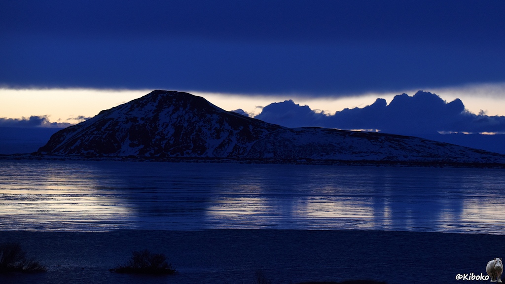 Ein spitzer Berg steht hinter einem eisbedeckten See. Wolken, Schatten und Ufer leuchten in dunklem Blau.