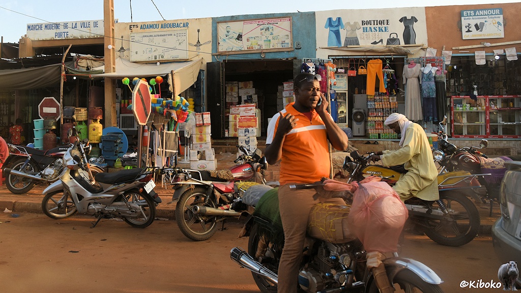 Das Bild zeigt im Vorergrund einen Mann im orangen Hemd auf einem Motorrad beim Telefonieren. Der Scheinwerfer und Lenker ist durch rosa Plastiktüten geschützt. Während die Tank und Sitzbank durch Pappe und Decken geschützt sind. Im Hintergrund sind Geschäfte in Garagengröße und fünf parkende Motorräder am Straßenrand.