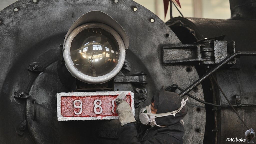Das Bild zeigt einen Mann mit Atemschnutzmaske, der die weißen Ziffern des Nummernschild 988 von roter Farbe befreit.