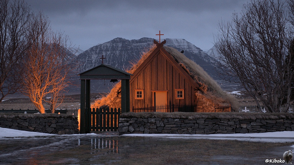 Eine kleine braune Holzkirche wird in der Abenddämmerung angestrahlt. Das Dach besteht aus gelbbraunem Gras und zieht sich bis zum Boden herunter. Nur der Giebel ist aus braunem Holz. Vor der Kirche ist eine Natursteinmauer. Eine Holzpforte mit einem Torbogen unterbricht die Steinmauer. Im Torbogen hängt eine Glocke.