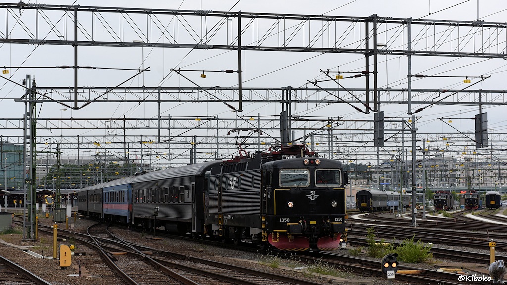 Das Bild zeigt eine schwarze, vierachsige Elektrolokomotive mit rotem Schneeräumer und gelben Griffstangen. Sie zieht einen Zug aus vier Schnellzugwagen. Der 2. Wagen ist blau lackiert und hat einen roten Streifen unter den Fenstern. Die anderen Wagen sind schwarz.
