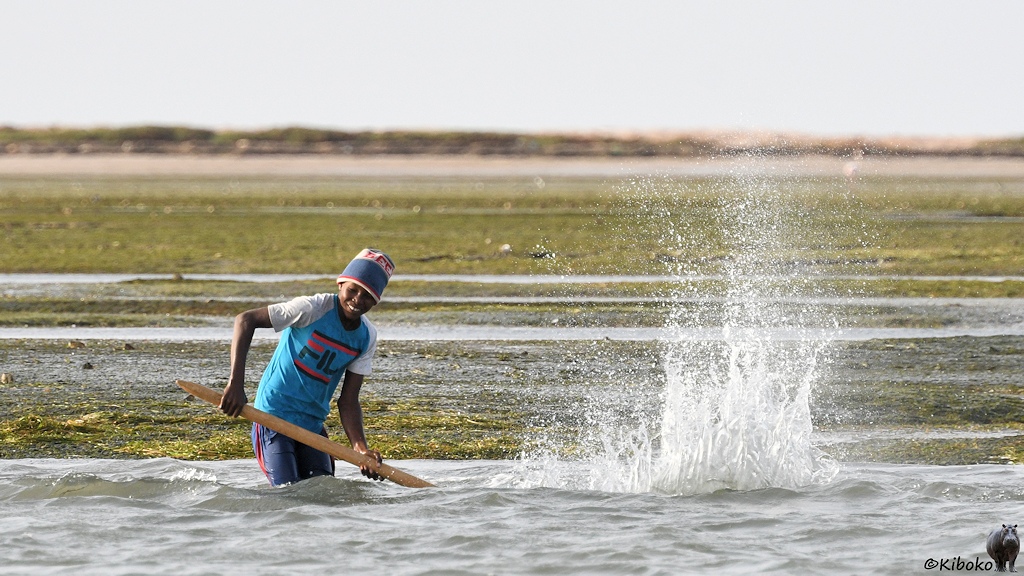 Das Bild zeigt einen Mann mit hellblauem T-Shirt, dunkelblauer Hose und Mütze mit den Beinen im Wasser stehen. Mit der langen Holzstange hat er auf das Wasser gescöagen. Eine Fontäne spritzt hoch. Im Hingergrund ist eine flache, grüne Insel.