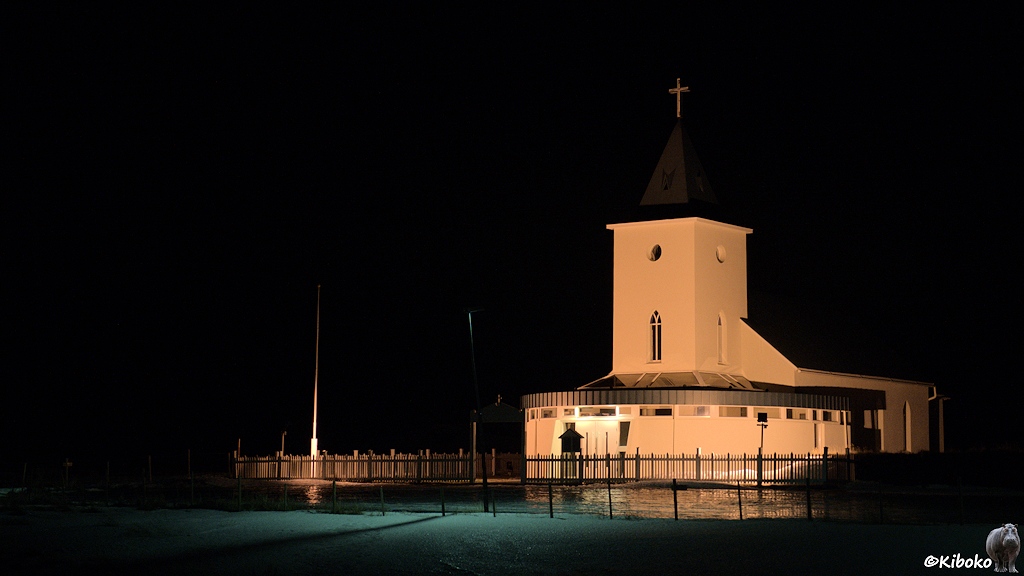Eine weiße Kirche mit einem viereckigen Turm wird nachts angestrahlt. Die Kirche ist durch einen Lattenzaun umringt.