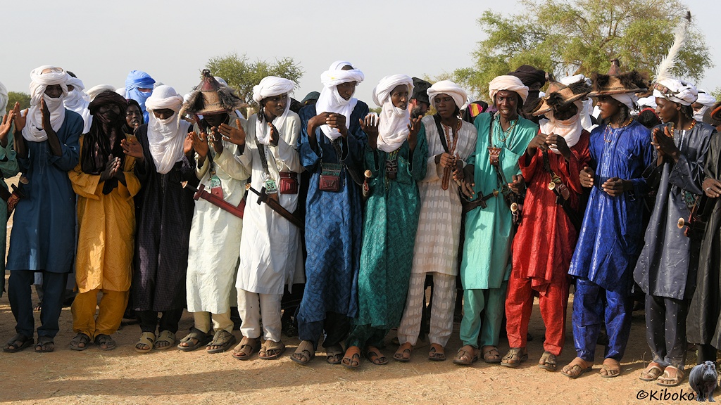 Das Bild zeigt eine tanzende Männerreihe überweigend mit weißem Turbanen und in bunten Gewändern in gelb, grün, blau, rot, weiß und grau.