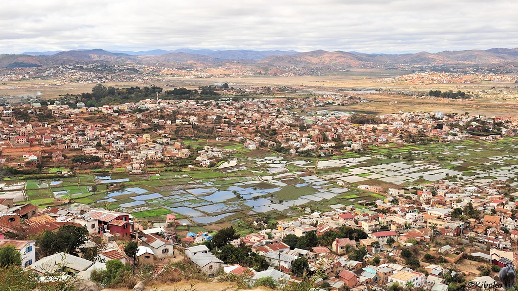 Das Bild zeigt eine Stadt von einem Berg. Im Vordergrund und auf einem gegenüberliegenden Hügel stehen ein- und zweigeschossige Steinhäuser. Im Tal sind Reisfelder die unter Wasser stehen. Im Hinergrund sind weitere Hügel auf denen ebenfalls Häuser stehen.