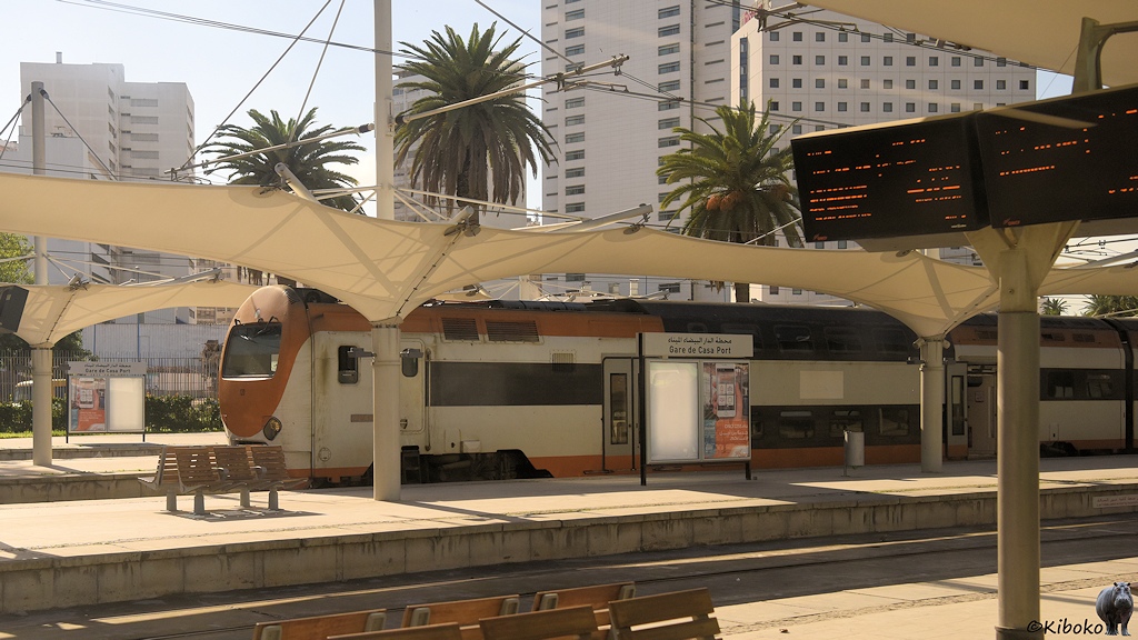 Das Bild zeigt einen weißen Doppelstocktriebwagen mit orangen Streifen und oranger Front hinter einen Bahnsteig mit einer weißen zeltförmigen Überdachung.