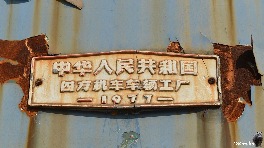 Das Bild zeigt ein weißes Fabrikschild mit zwei Reihen chinesischer Schriftzeichen. Darunter ist das Baujahr: 1977. Das Schild ist schon in der Mitte gerissen. Die Lok dahinter hat große Rostflecken und die hellblaue Farbe blättert ab.