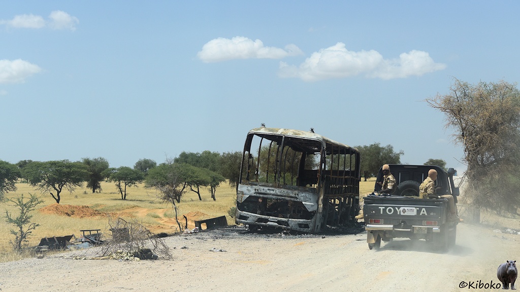 Das Bild zeigt ausgebrannten Reisebus am Rand einer staugigen Straße. Das Buswrack passiert eine grüner Geländewagen mit zwei Soldaten auf der Ladefläche.