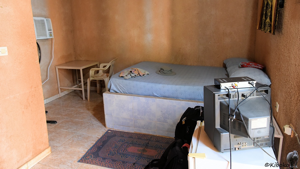 Das Bild zeigt das Zimmer vorn Kiboko. Hinten ist das Bett mit hellblauem Bezug der Matratze, die auf einen Betonsockel liegt. Am Fußende ist eine Klimaanlage an der Wand montiert. Im Vordergrund steht ein Röhrenfernseher im silbergrauen Gehäuse. Die Wände sind terrakottafarben.