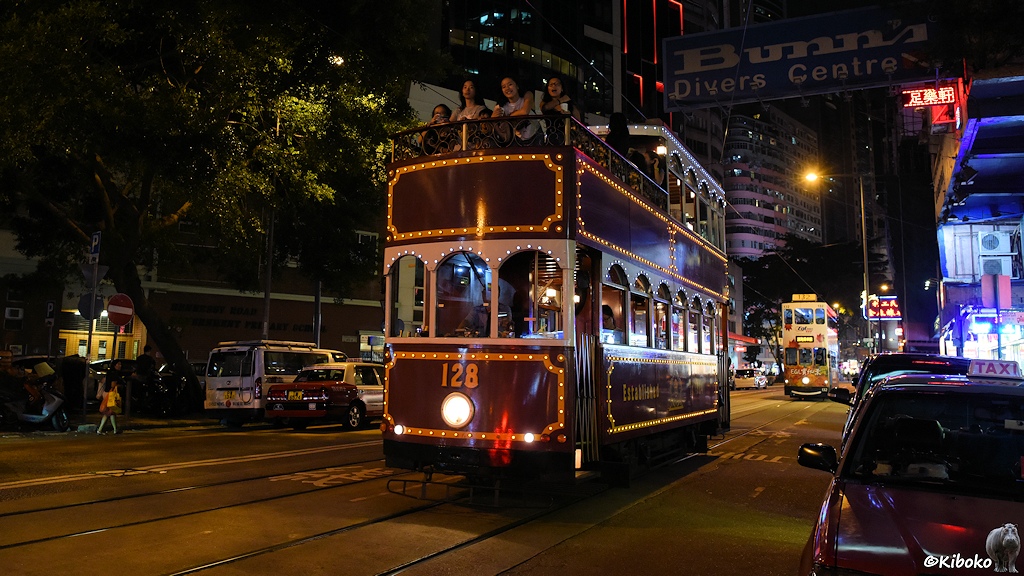 Das Bild zeigt die rot-weiße Straßenbahn 128 mit offenen Oberdeck auf Sonderfahrt. Die Straßenbahn ist mit Bändern von kleinen Lämpchen dekoriert.