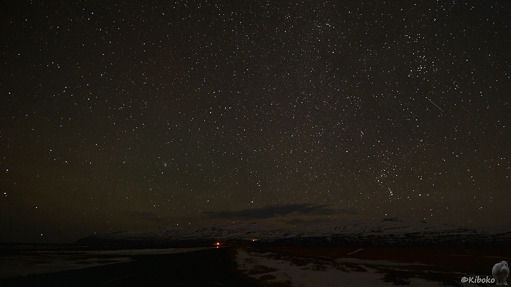 Ein Sternenhimmel leuchtet über eine dunkle Straße, die auf eine schneebedeckte Bergkette führt. Im Sternenhimmel sind ein paar Punktlinien und Striche von Flugzeugen und Satelliten.