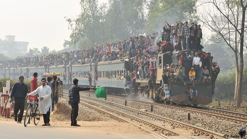 Das Bild zeigt einen übervollen Zug in einer leichten Kurve. Auf der linken Seite zeigt ein Mann eine grüne Fahne am ausgestreckten, waagerechten Arm. Die blau-beige Diesellokomotive ist unter den Menschmassen kaum zu sehen. Sogar vorn auf der Kupplung sitzt ein junger Mann. Auf dem Dach ist eine Wand von Menschen. Aus jedem Fenster schauen Köpfe heraus. An jedem Trittbrett hängt eine Menschentraube.