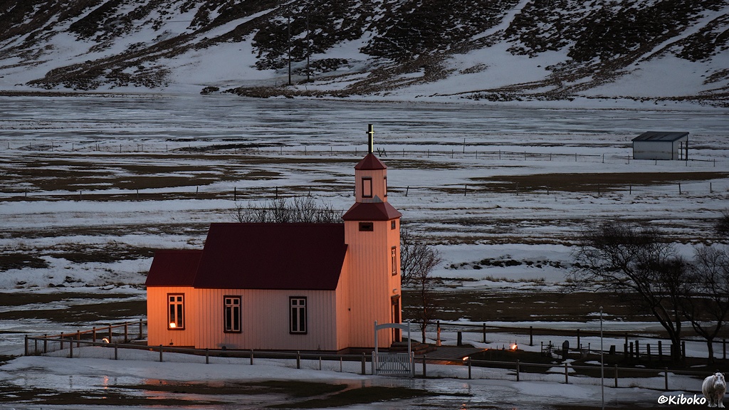 Kleine weiße Holzkirche, die durch gelbes Licht angestrahlt wird. Im Hintergrund sind schneebedeckte Wiesen.