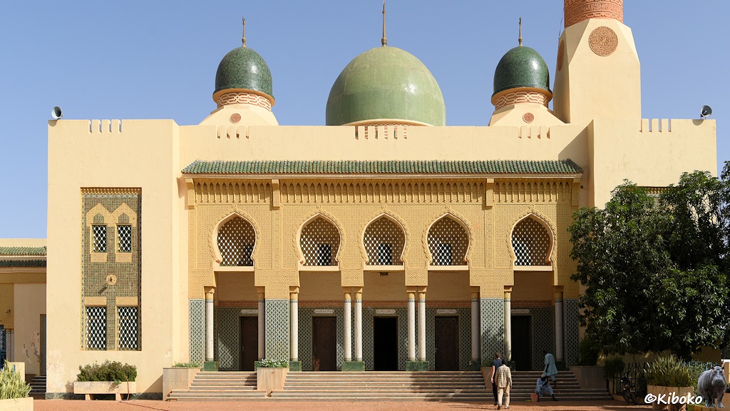 Das Bild zeigt die beige und gelbe Fassade der großen Moschee. Eine breite Treppe mit 10 Stufen führt zu 5 Türen. Dazwischen sind weiße Säulen die orientalische Ornamente halten. Ein Vordach mit grünen Ziegeln schließt den Eingangsbereich ab. Auf dem Dach sind drei grüne Kuppeln. Eine größere in der Mittel und zwei kleinere am Rand.