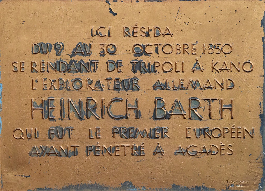 Das Bild zeigt eine golden lackierte Metalltafel mit teilweise abgeblätterten Farbe mit der Aufschrift: ICI RÈSIDA, Du 9 au 30 Octobre 1850 se rendant de Tripoli a Kano L'Explorateur Allemand HEINRICH BARTH qui fut le premier Euroüéen Ayant Pénetré Européen. Ayant Pénetré À Agadès.
