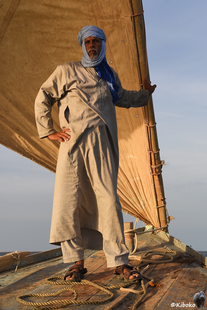 Das Bild zeigt eine Hochformataufnahme eines stehenden Mannes in der Morgensonne. Er trägt einen hellblauen Turban und ein beigefarbenes Gewand. Hinter ihm ist das aufgeblähte Segel. Eine Hand greift um den Mast. Die andere Hand ist in die Hüfte gestützt.