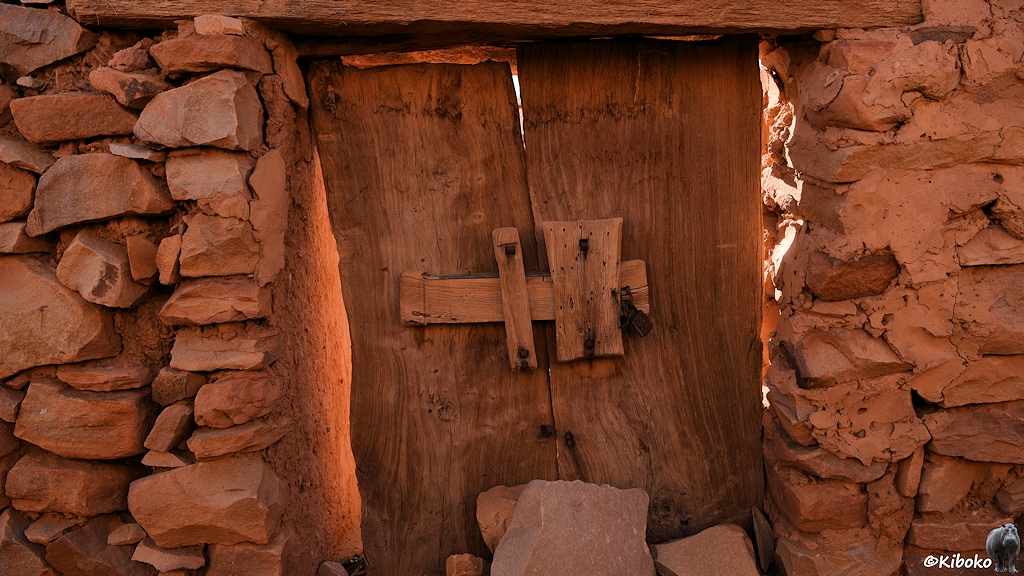 Das Bild zeigt eine Holztür aus zwei senkrechten massiven Brettern in einer rötlichen Natursteinwand. Breite Holzriegel und ein verrostetes Vorhängeschloss sichern die Tür.