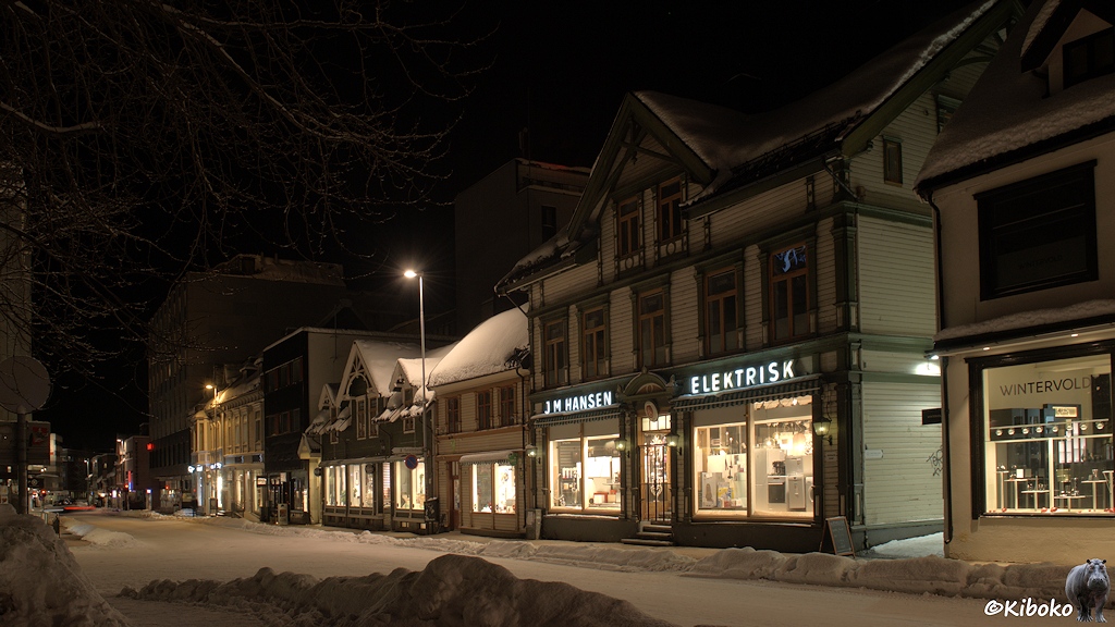 Das Bild zeigt eine verschneite Straße mit einer Zeile zweistöckiger Holzhäuser. Im Ergeschoss sind beleuchtete Geschäfte. Einige Geschäfte haben Leuchtschriften über den Schaufenstern.