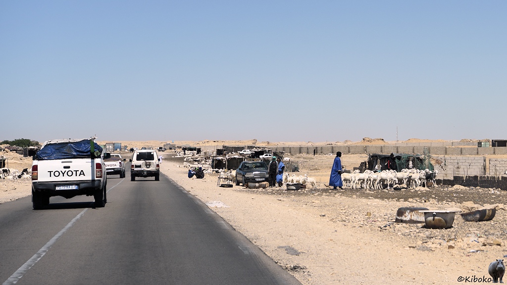 Das Bild zeigt eine zweispurige Teerstraße auf der gerade Geländewagen sich überholen. Am rechten Bildrand sind Schafherden, dunkle Schuppen, Männer in blauen Gewändern, ein Mercedes und drei Badewannen.