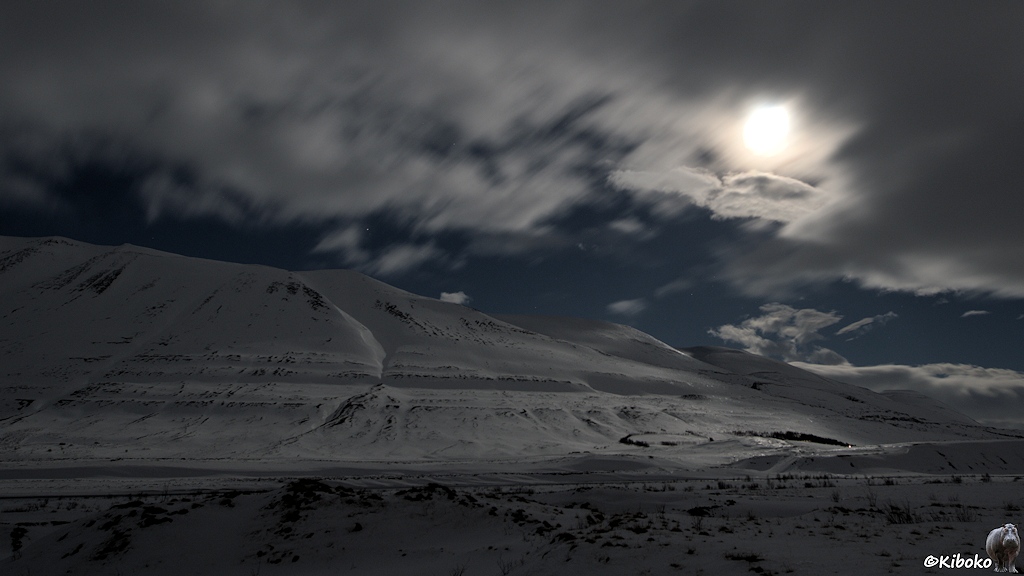 Der Mond leuchtet diffus durch eine Wolkendecke. In einer Wolkenlücke leuchten die Sterne über einer schneebedeckten Landschaft