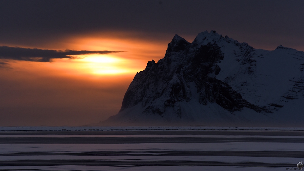 Das Bild ist ähnlich dem vorhergehenden Bild. Die Sonne ist dem Bergmassiv näher gekommen und taucht die zugefrorene Bucht in ein orangefarbenes Licht.