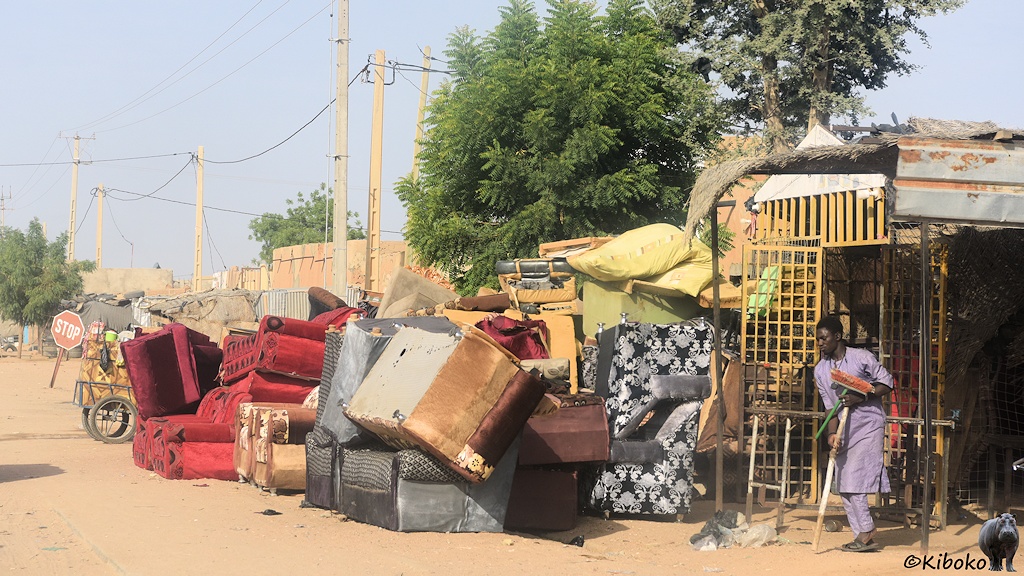 Das Bild zeigt Sofas und Polstersessel in grau, braun, rot und hellgrün am rechten Straßenrand auf hellbraunem Sand. Sie sind teilweise kopfüber gestapelt. Am rechten Bildrand ist ein Ladenschgeschäft aus einem gelben Verschlag. Darin steht ein Mann in violetter Kleidung und hällt einen Besen mit dem Stil nach unten.